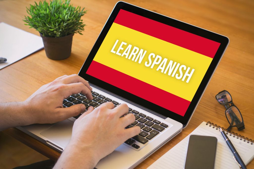 człowiek uczy się hiszpańskiego i trzyma dłonie na laptopie,  laptop leży na drewnianym biurku i ma napis na ekranie learn spanish, obok leży notes, długopis, telefon i okulary