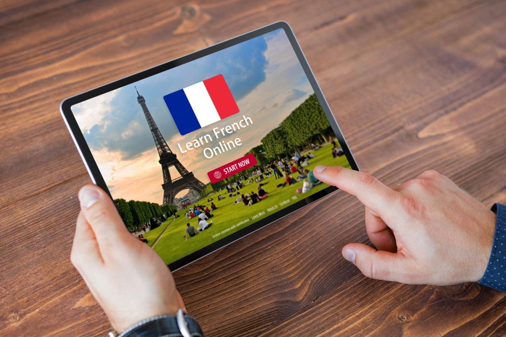 dłonie trzymają tablet z nauką francuskiego, widać w nim wieżę eiffla, flagę Francji, napis start, zielone miejsce we Francji, natomiast w tle widać drewniany stół