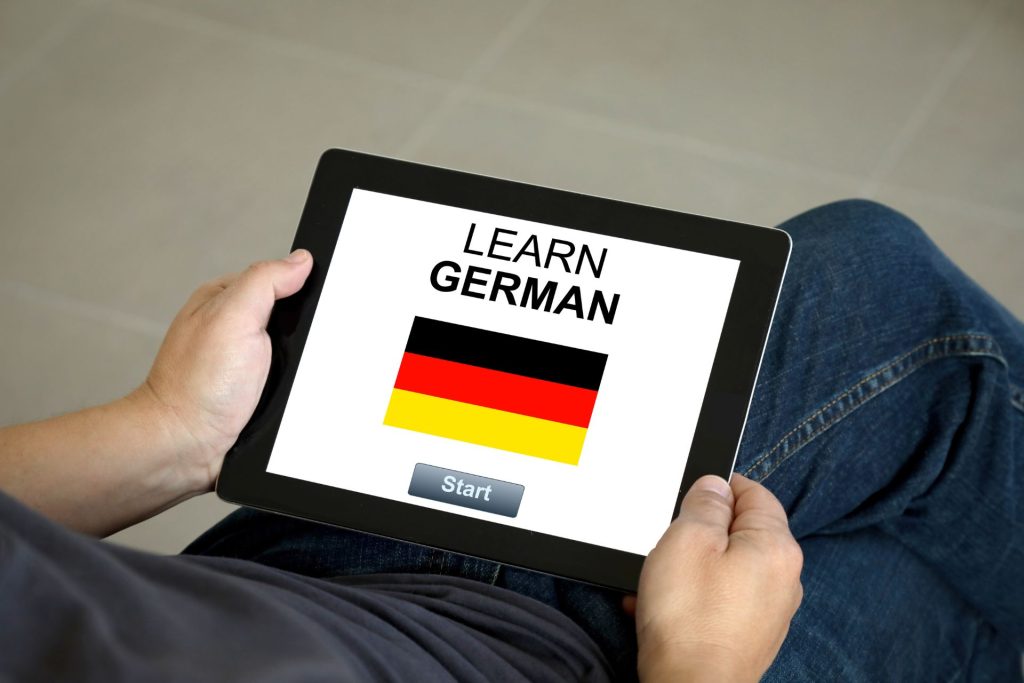 mezczyzna ma lekcje nauki niemieckiego online, w obu rękach trzyma tablet, w tle widać płytki na podłodze