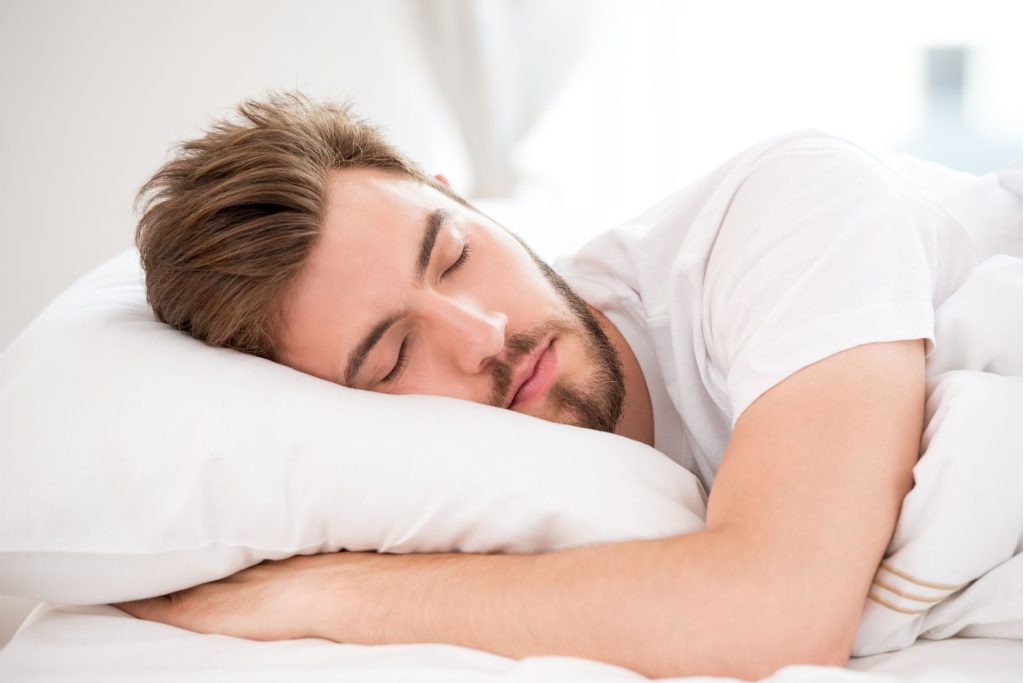 mężczyzna z brodą w białej koszulce śpi na łóżku i na białej poduszce