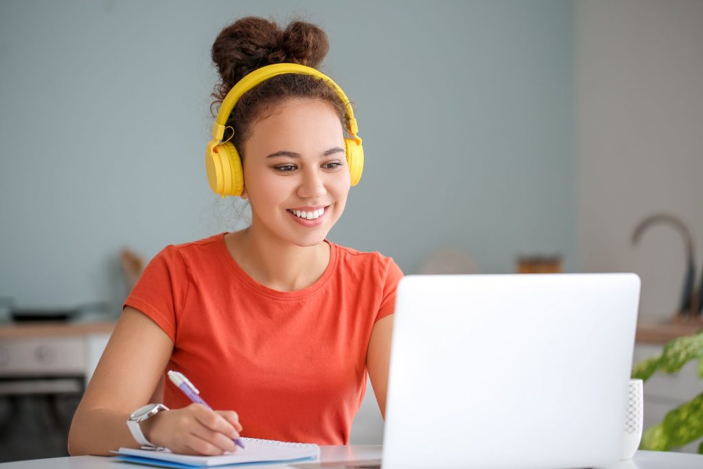 Eine Frau mit Kopfhörern auf den Ohren und einem Stift in der Hand blickt auf einen Laptop und lernt polnishe Wörter für den Urlaub